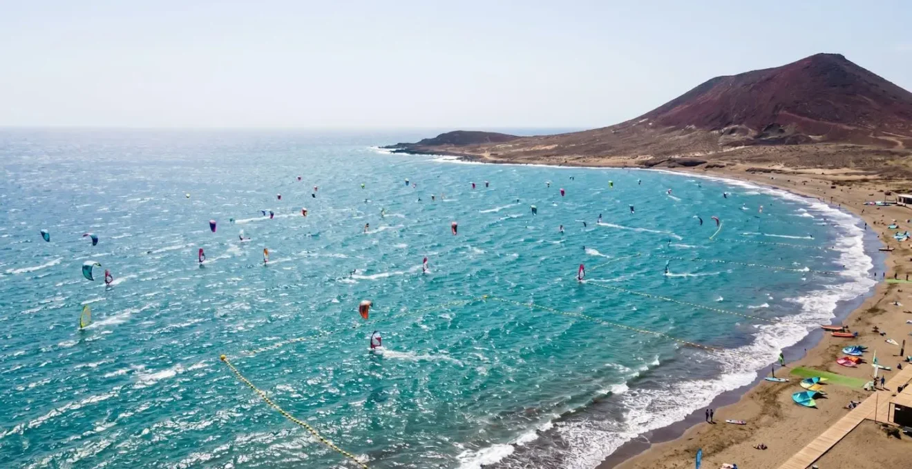 Aerial view of El Médano Bay showing kiteboarders and windsurfers navigating choppy waters with yellow safety buoys and Montana Roja in the background