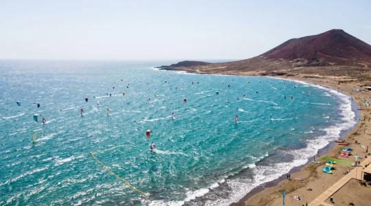 Aerial view of El Médano Bay showing kiteboarders and windsurfers navigating choppy waters with yellow safety buoys and Montana Roja in the background