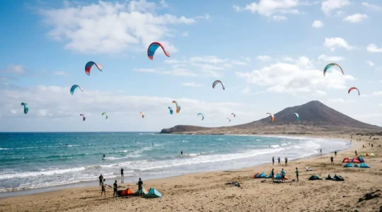 Wide panoramic view of El Médano bay with colorful kites in stable trade winds, volcanic Montaña Roja in background, sandy beach and turquoise waters ideal for beginner kitesurfers