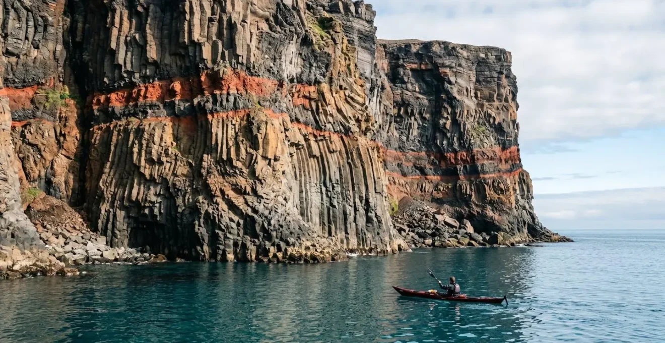 Sea kayaker observing layered volcanic basalt cliffs with columnar jointing and red paleosol layers from water perspective