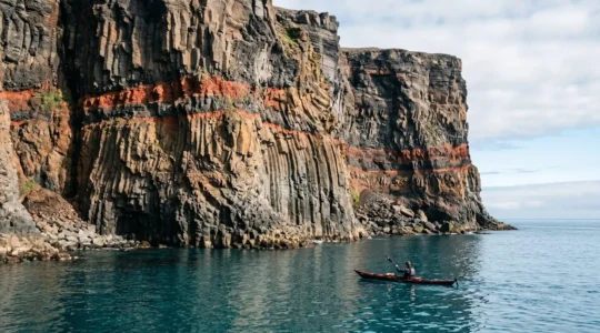 Sea kayaker observing layered volcanic basalt cliffs with columnar jointing and red paleosol layers from water perspective