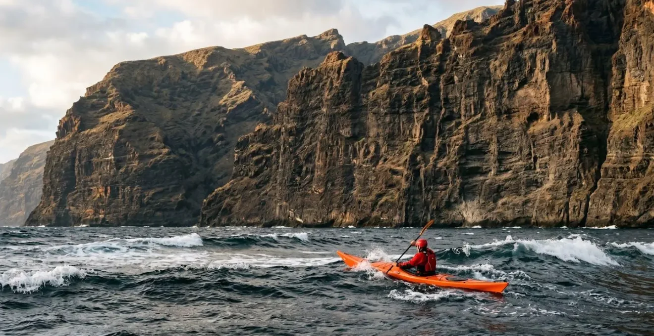 Sea kayaker paddling against strong trade winds along dramatic volcanic cliffs of Los Gigantes in Tenerife, with towering rock walls and choppy Atlantic waters