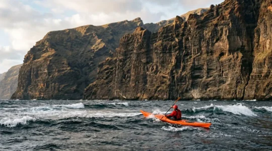 Sea kayaker paddling against strong trade winds along dramatic volcanic cliffs of Los Gigantes in Tenerife, with towering rock walls and choppy Atlantic waters