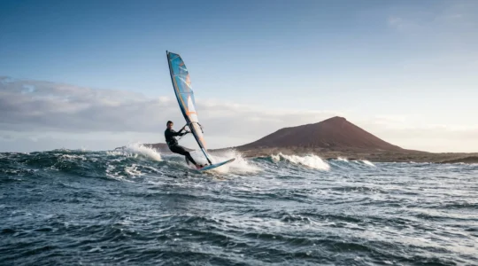 Windsurfer navigating choppy Atlantic swell in El Médano with colorful sail against volcanic landscape backdrop